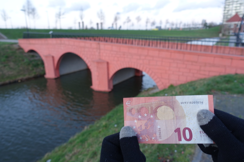 foreground: a euro note. background: the bridge from that note